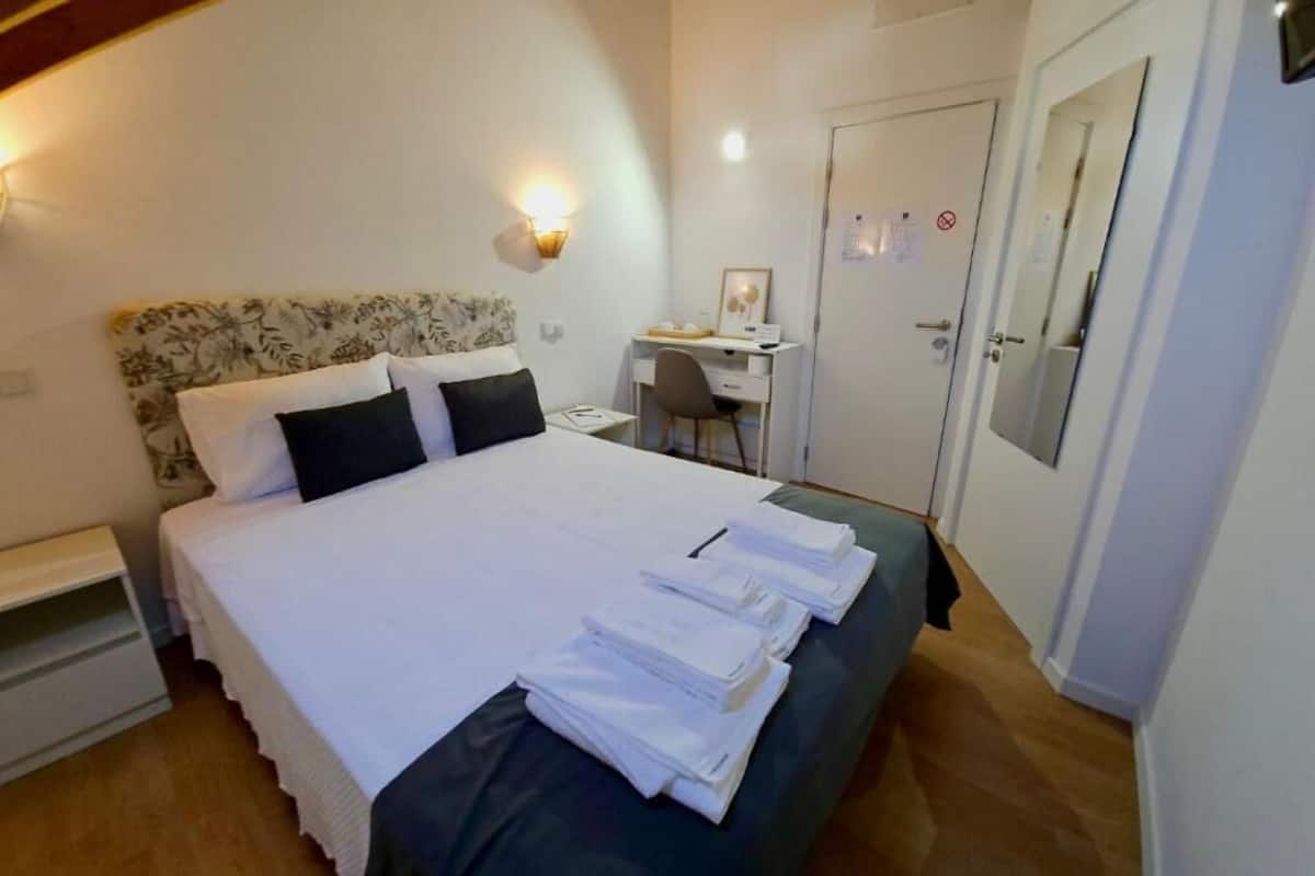 Un lit bien fait, avec du linge de lit blanc, un bureau avec une chaise, une armoire et un luminaire fixé au mur.