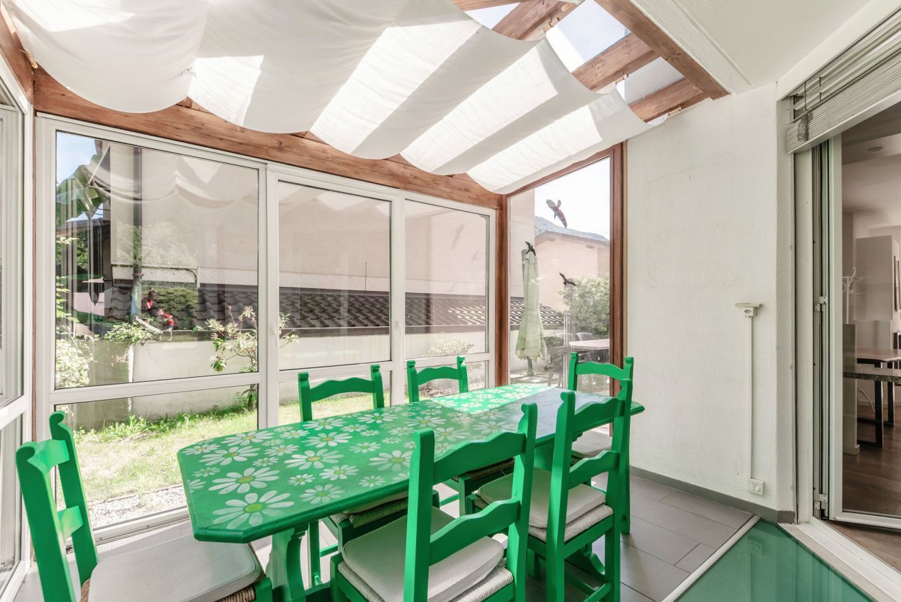Une terrasse couverte avec une table et des chaises, de grandes fenêtres et une vue sur l’extérieur.