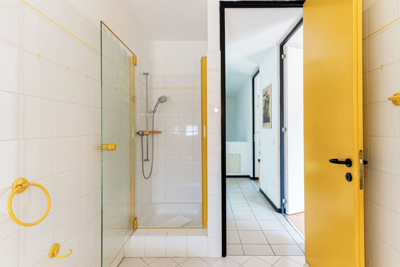 A modern bathroom with a glass-enclosed shower, white tiled walls, and a yellow door.