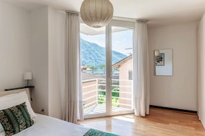 A hotel room with a bed, a nightstand, a lamp, a large window with curtains, and a view of a mountain. - Detached house with swimming pool (MOLINI POOL HOUSE) (Maroggia)