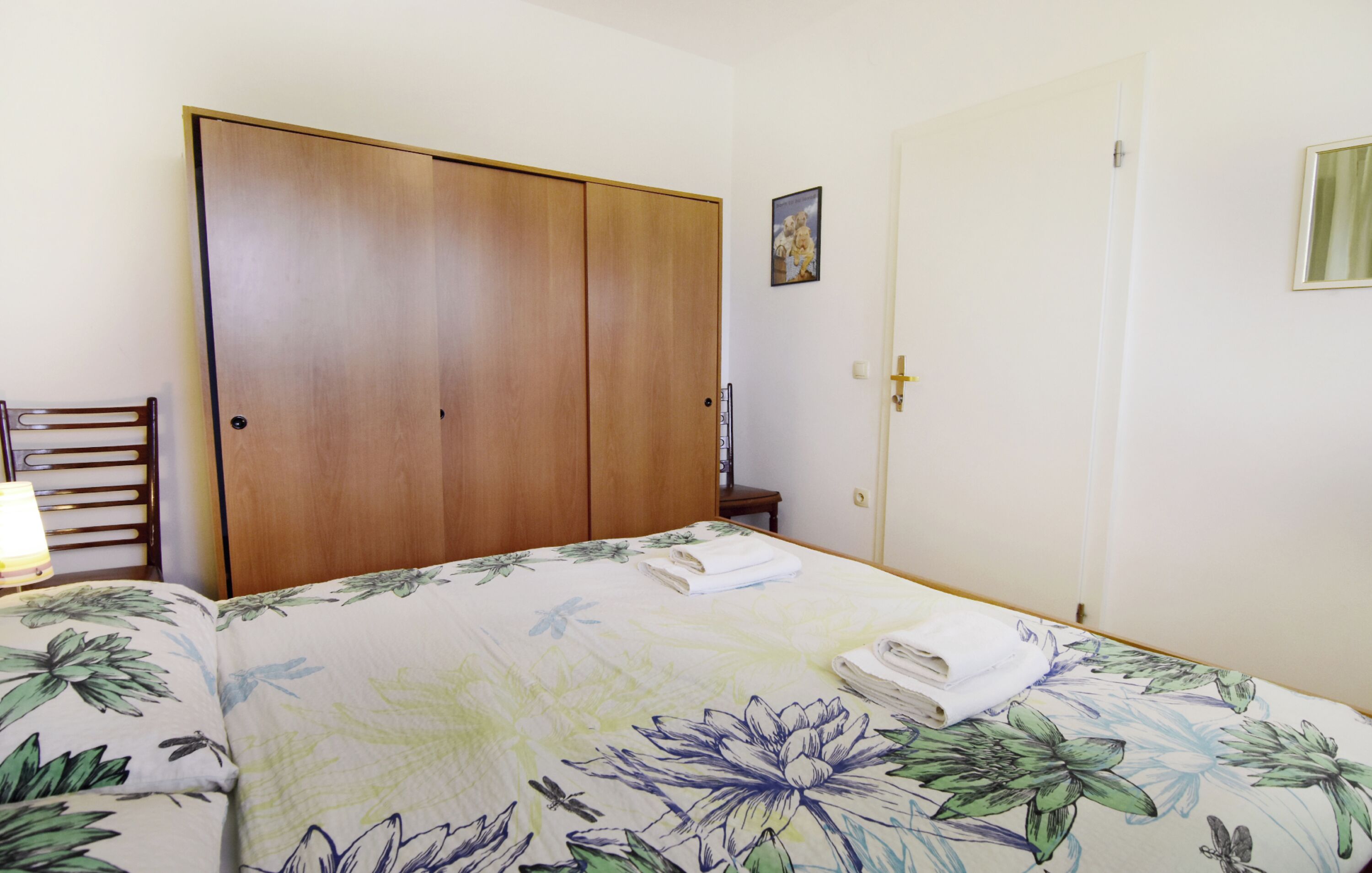 Een slaapkamer met een bed, een houten kledingkast, een deur en een kleine tafel met een lamp.