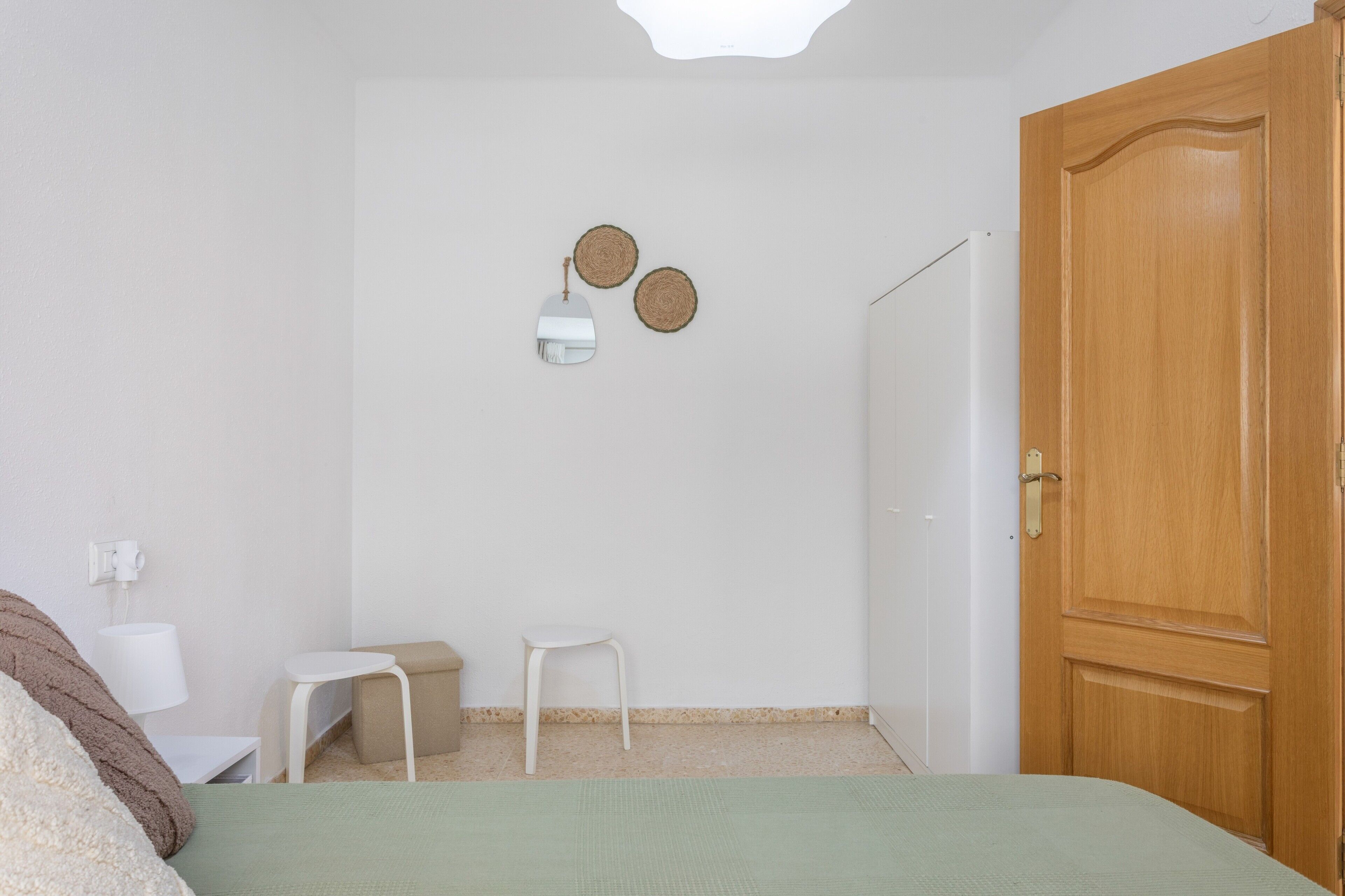 Una habitación con cama, puerta de madera, pared blanca, pequeña mesa blanca, banco beige y lámpara fijada a la pared.
