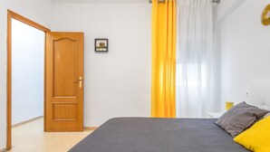 A bedroom with a wooden door, a bed with grey and yellow bedding, a small framed picture on the wall, and a window with yellow curtains. - Apartment 'Playa Valencia' with Terrace, Wi-Fi and Air Conditioning (Mareny de Barraquetes)