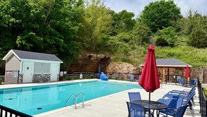 Una piscina con lettini e ombrelloni, circondata da alberi e un piccolo edificio.