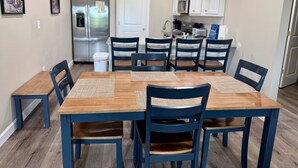 Zona pranzo con tavolo di legno e sedie blu, cucina sullo sfondo e quadri appesi al muro.