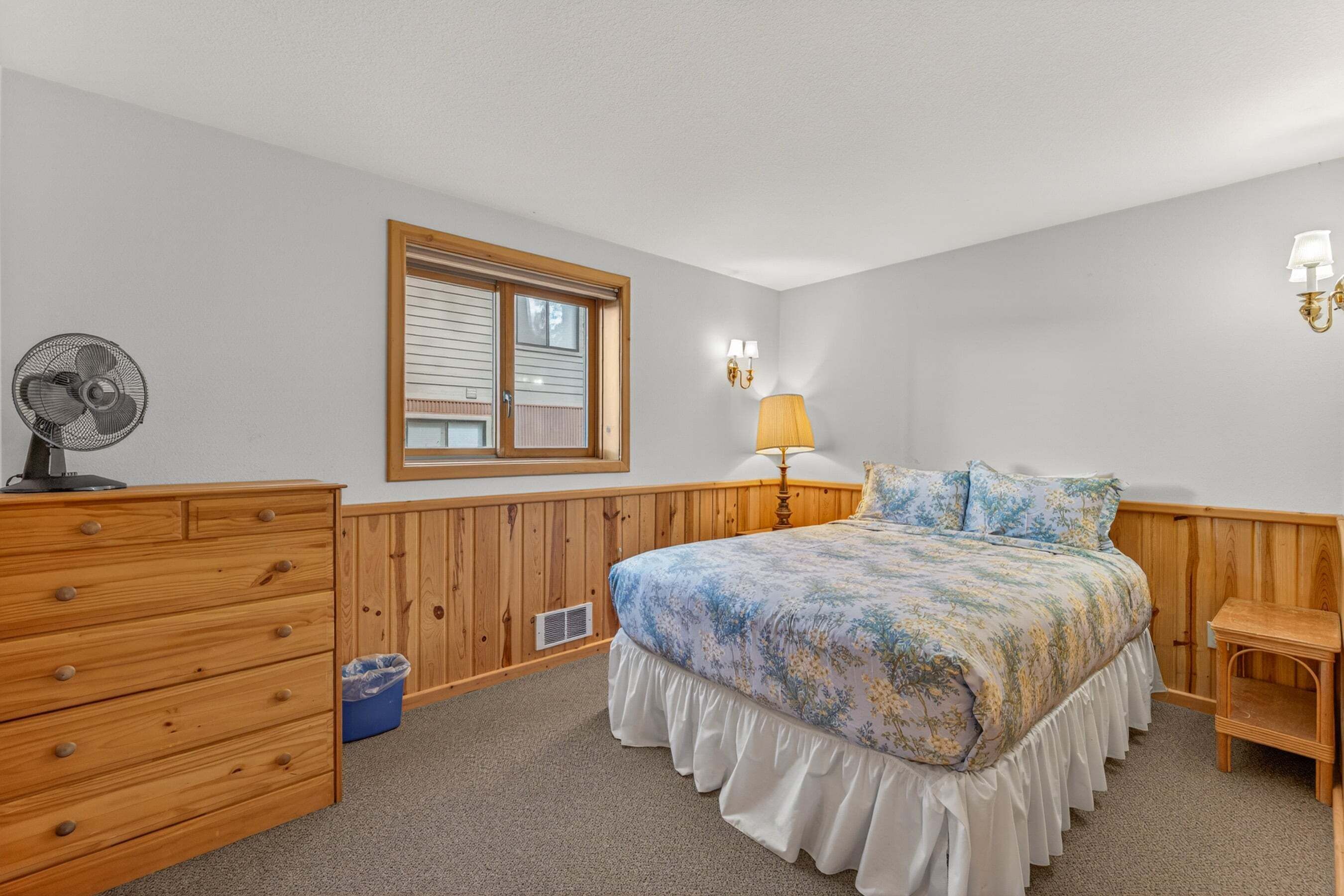 Ein Schlafzimmer mit Bett, Kommode, Fenster, Lampe und Ventilator.