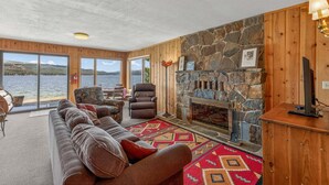 L’intérieur d’un chalet avec une cheminée en pierre, des murs en bois, un grand canapé et une vue sur le lac.