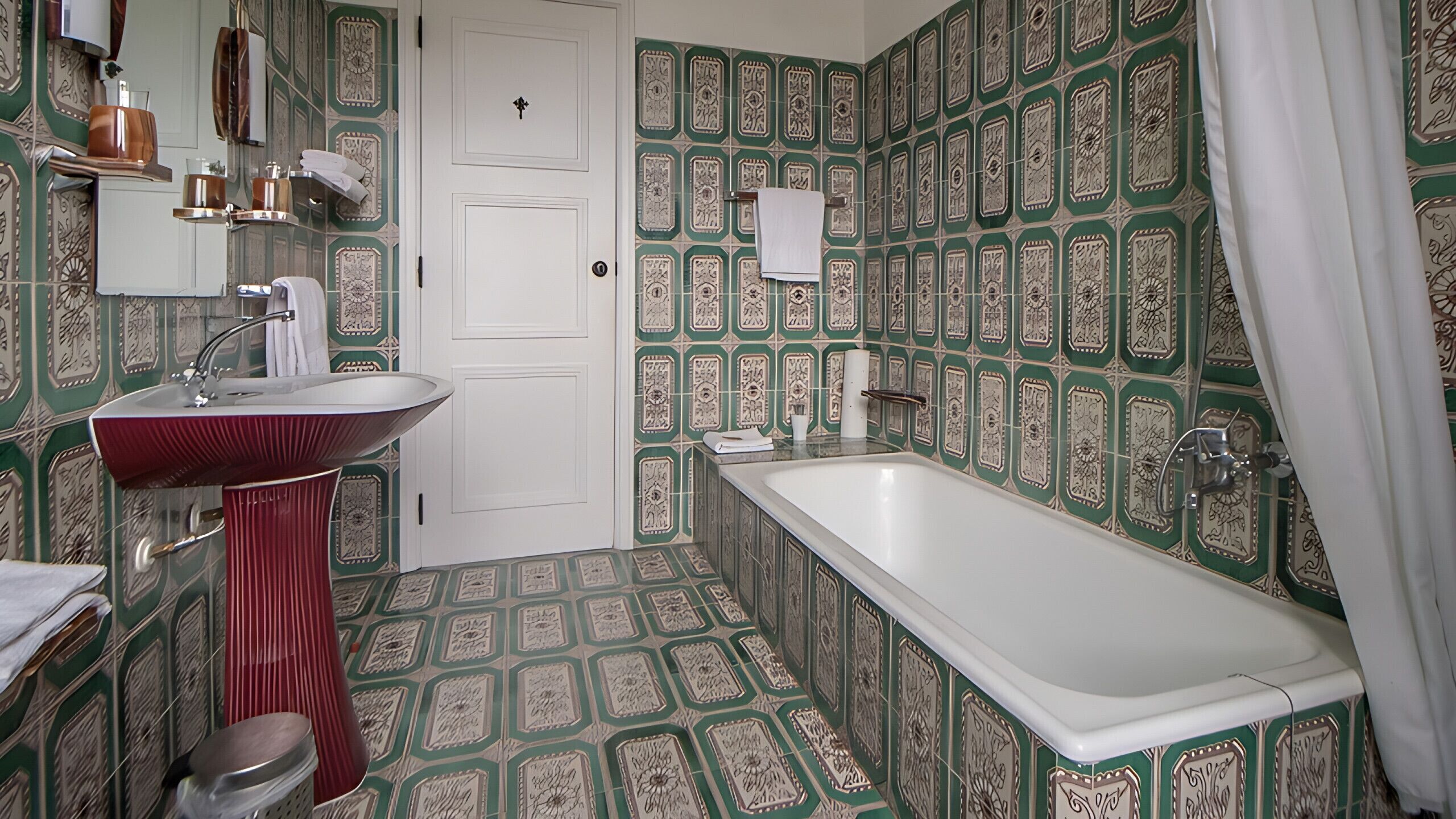 Ein Badezimmer mit gemusterten Fliesen, einer weißen Badewanne, einem roten Waschbecken und einer Tür.