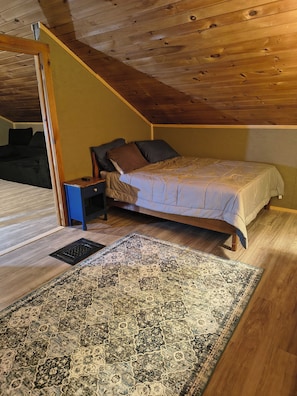 Ein Schlafzimmer mit Holzbalkendecke, einem Bett, einem Nachttisch, einem Sessel und einem gemusterten Teppich.