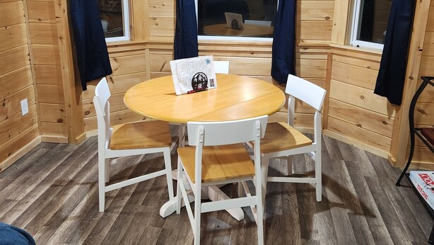 Een houten blokhutinterieur met een ronde houten tafel en witte stoelen, grote ramen met gordijnen en een houten vloer.