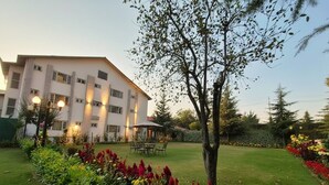 A building with a flat roof, multiple windows, and a well-maintained garden with various plants and a tree. - Hotel Meridien River Front Rajbagh (Srinagar)