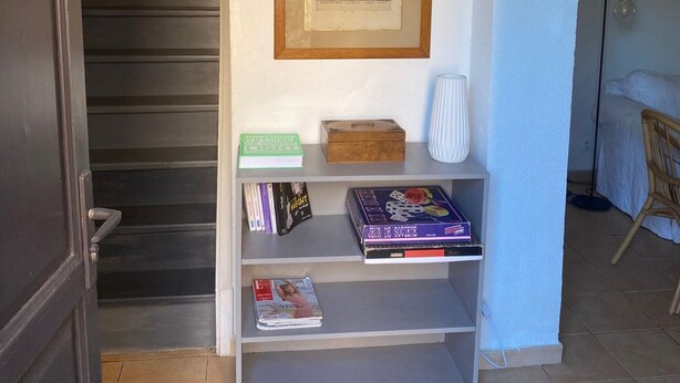 A hallway with a staircase, a framed picture on the wall, a bookshelf with books and a board game, and a bed in the adjacent room. - Villa Spacieuse Avec Piscine Priv e 150 m de la Plage Calvi Centre (Calvi)