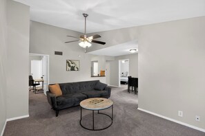 Ein Wohnzimmer mit einem dunklen Sofa, einem runden Couchtisch, einem Deckenventilator und einem Essbereich im Hintergrund.