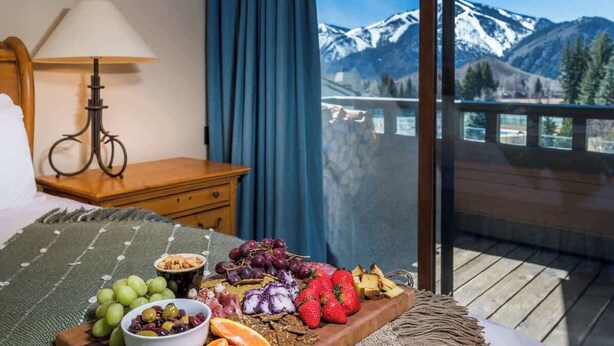 Ein Balkon mit Blick auf die Berge, ein Holztisch mit Obst und Käseplatte, eine Lampe und ein Bett mit grüner Decke.