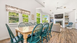A dining area with a wooden table and chairs, a fireplace, a television, and a view of greenery through large windows. - Sweet Caroline's: Flexible Stays, Dog-friendly, Close to Beach (Corolla)