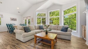 A spacious living room with a sectional sofa, a wooden coffee table, and large windows offering a view of greenery. - Sweet Caroline's: Flexible Stays, Dog-friendly, Close to Beach (Corolla)