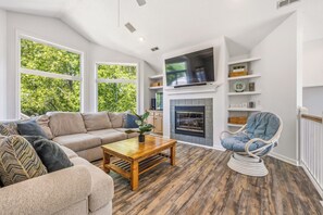 A living room with a sofa, armchair, coffee table, fireplace, TV, and large windows. - Sweet Caroline's: Flexible Stays, Dog-friendly, Close to Beach (Corolla)