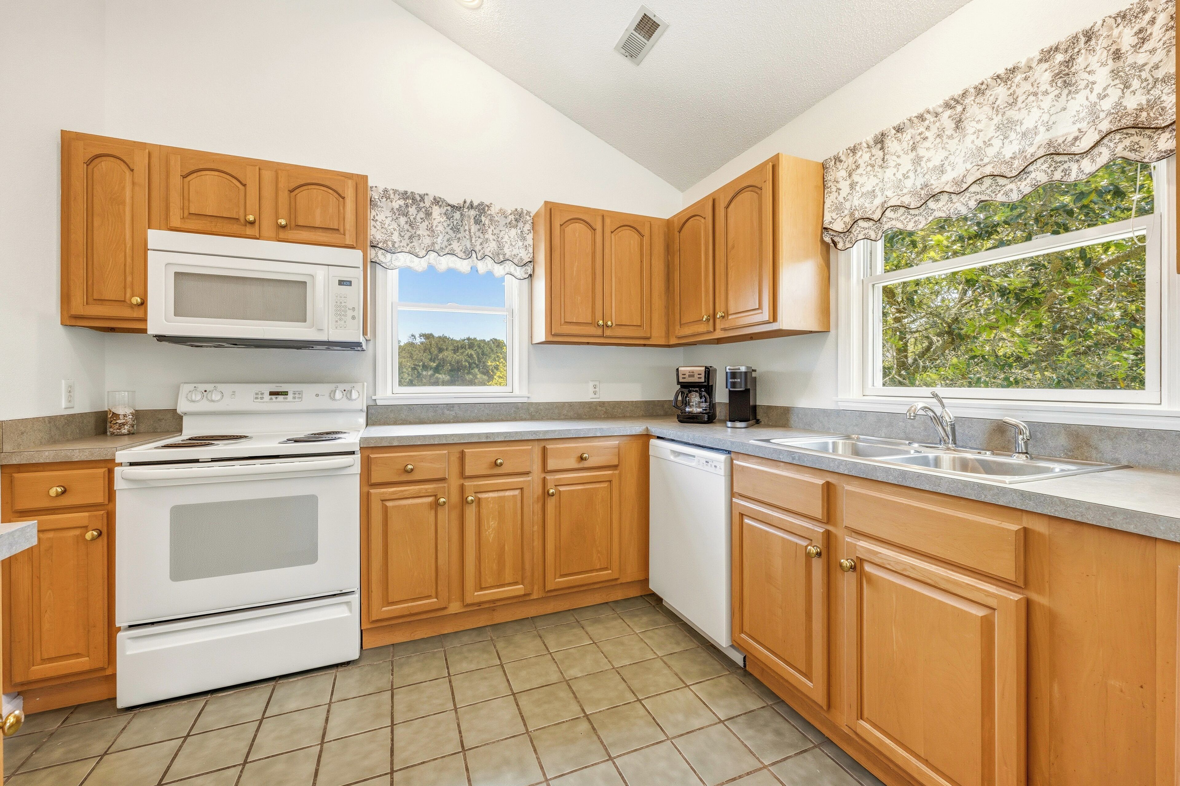 A kitchen with wooden cabinets, a microwave, stove, dishwasher, and a window with a view of trees.