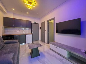 A modern living room with a sofa, a coffee table, a flat-screen TV mounted on the wall, a kitchen area with cabinets and a refrigerator, and a door leading to another room. - 13 - DELFINI Suites by SDR (Paralia)