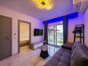 A modern living room with a sofa, a flat-screen TV, a small table, and a sliding glass door leading to a balcony. - 13 - DELFINI Suites by SDR (Paralia)