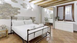 Une chambre avec un cadre de lit en métal, du linge de lit blanc, des éléments décoratifs en pierre, des poutres en bois, une commode blanche et une fenêtre avec vue.