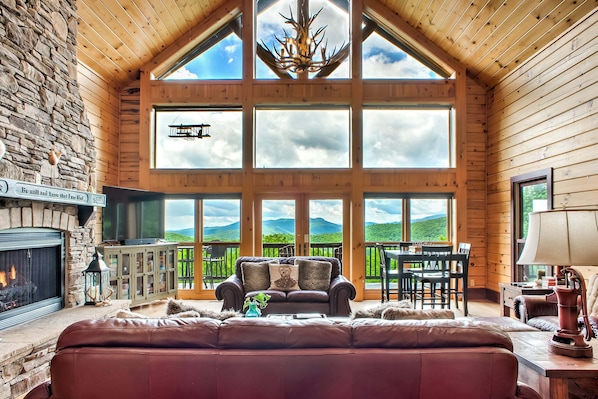 A cozy cabin interior with a stone fireplace, leather sofa, and a large window overlooking a scenic landscape.