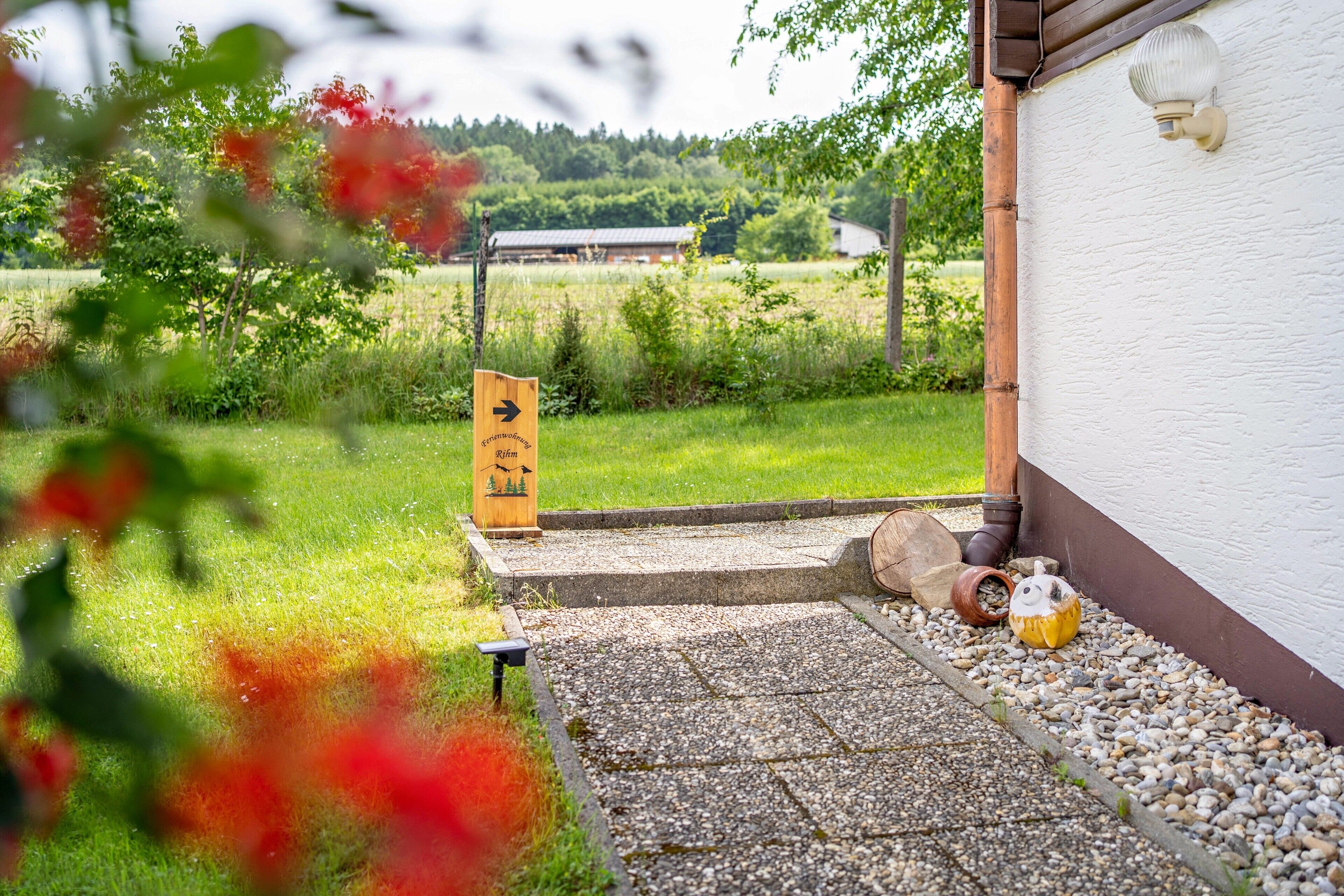 Un chemin mène à un bâtiment dont la façade est blanche, orné d’une enseigne en bois portant un symbole, et devant lequel se trouve un petit jardin avec des galets et une décoration en forme de citrouille.