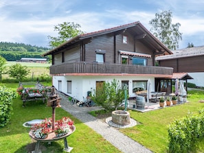 Een houten huis met twee verdiepingen, een balkon, een tuin met bloemen en een vuurkorf, en een heldere lucht.