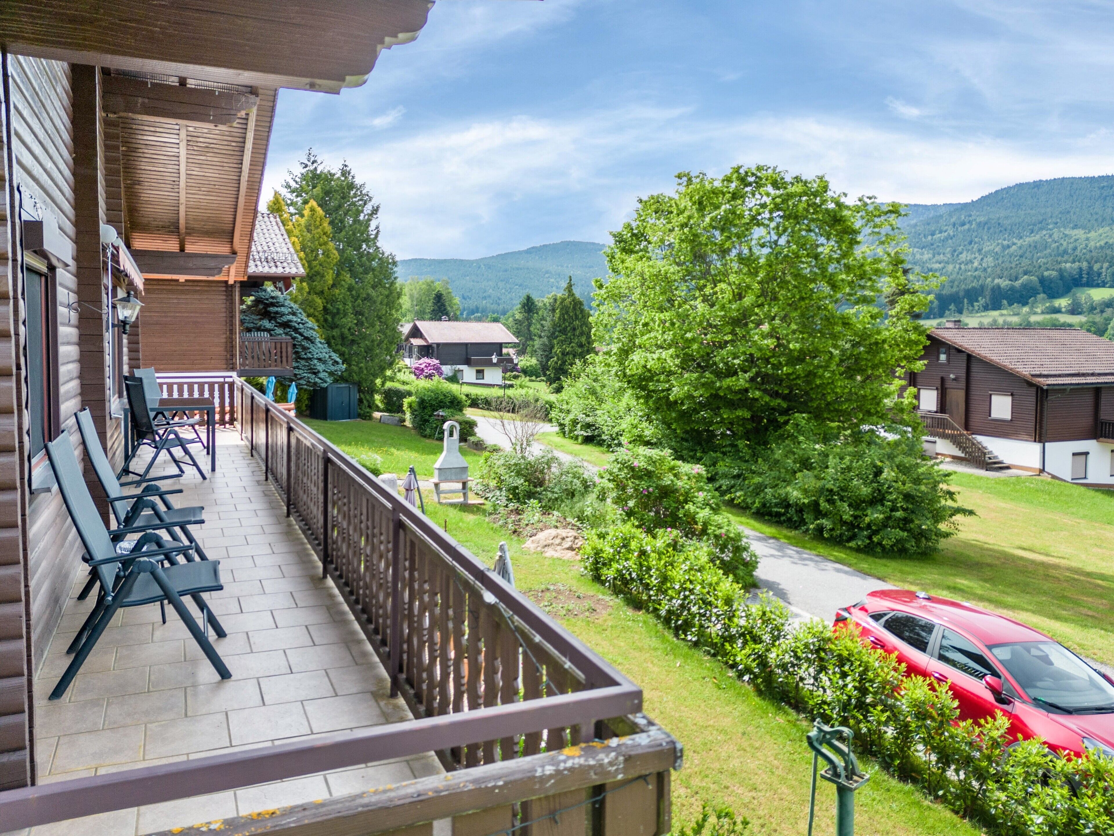 Un balcon avec des sièges d’extérieur, une voiture rouge garée à proximité, et une vue sur un paysage verdoyant avec des maisons et des collines.