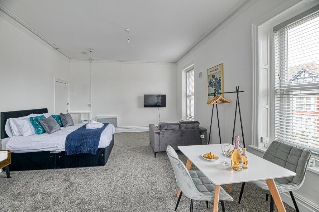 A hotel room with a bed, a gray sofa, a dining table with chairs, a TV, and a window with blinds.