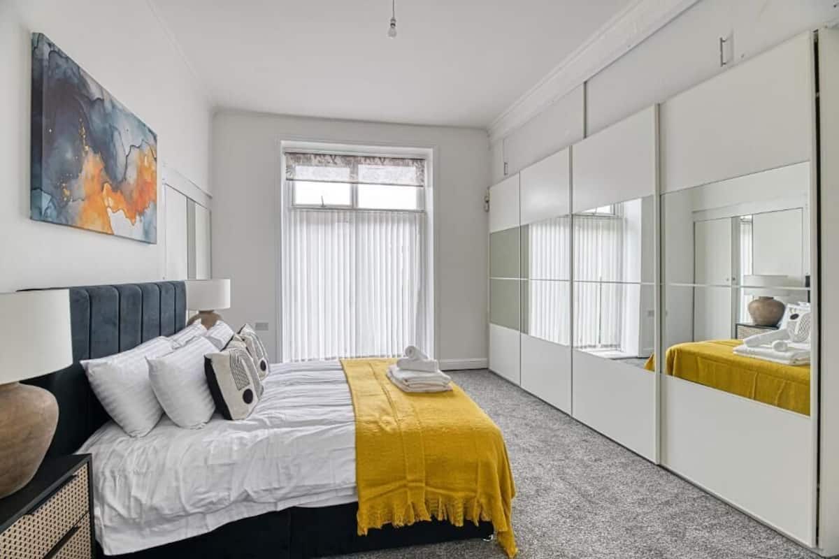 Un dormitorio con cama, mesita de noche, espejo y ventana con persianas.