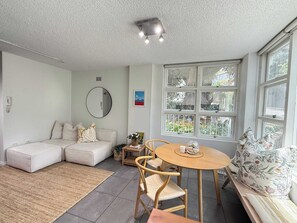 A living room with a sofa, a round table, and a window with a view of greenery. - Chic Waterfront Stay with Prime Sydney Access (Lavender Bay)