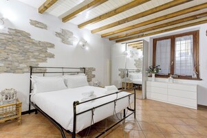 Un dormitorio con estructura de cama metálica, ropa de cama blanca, detalles de pared de piedra, techo de madera y una ventana con cortinas.