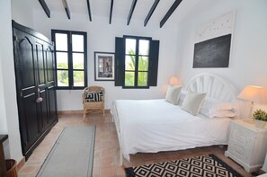 A bedroom with a bed, two windows, a chair, a nightstand, and a wall-mounted picture.