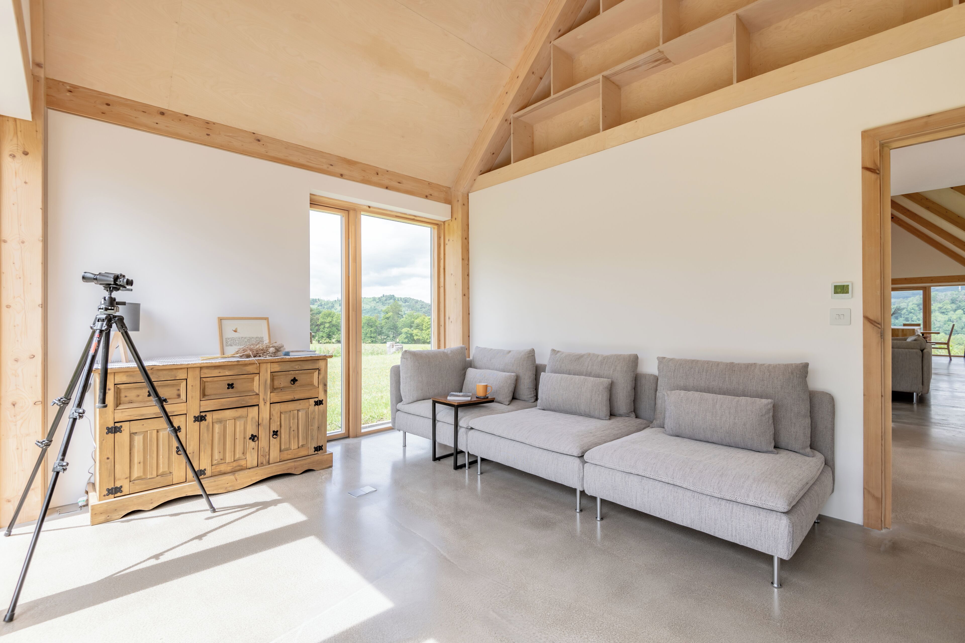 A spacious room with a large window, a wooden ceiling, a grey sofa, and a tripod.