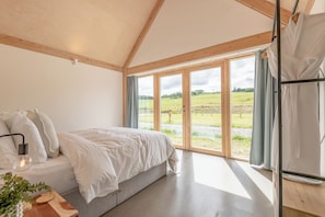 A bedroom with a bed, a bedside table with a lamp, a window with curtains, and a view of the outdoors.