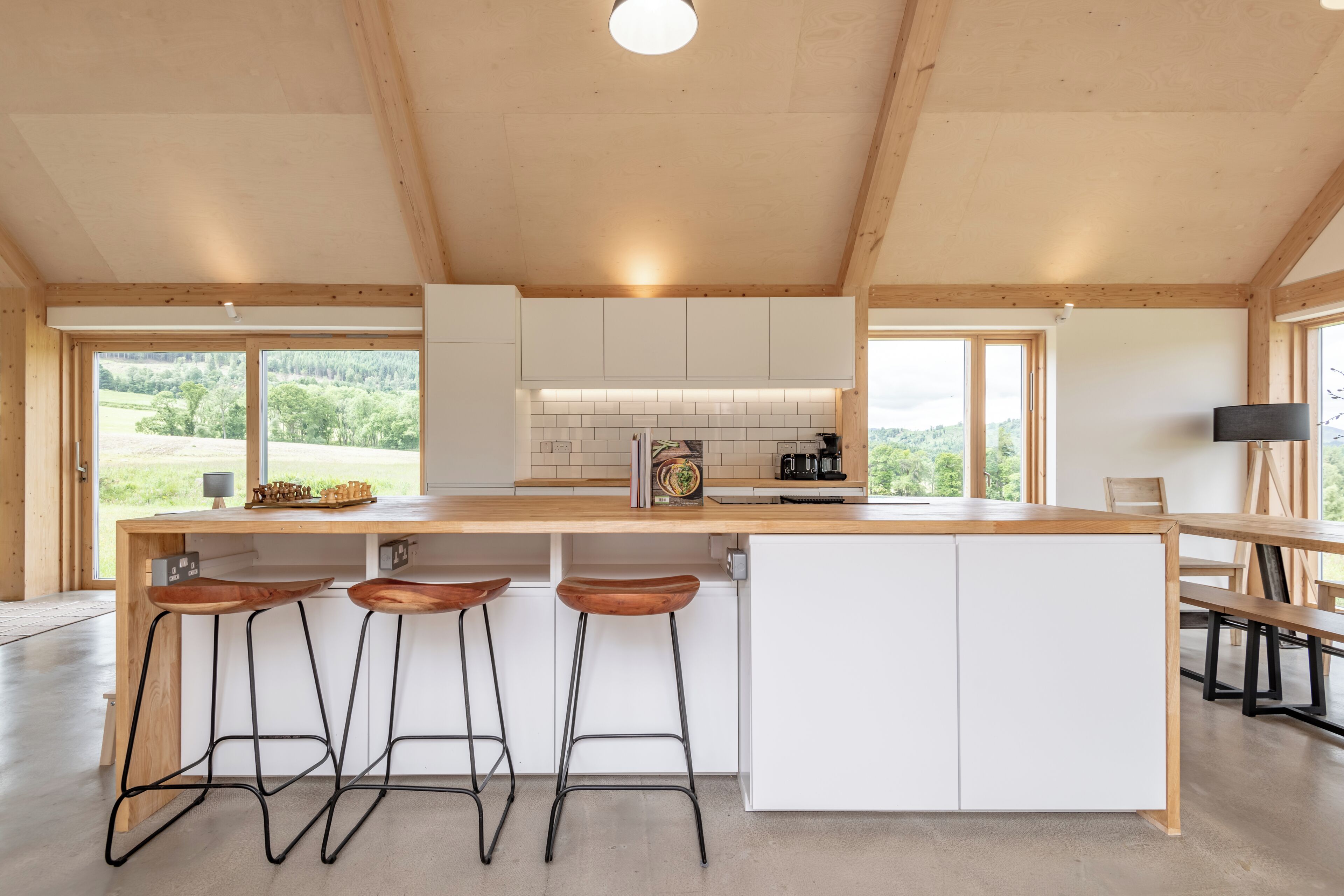 A modern kitchen with wooden beams, white cabinets, a large island with stools, and large windows offering a view of greenery.