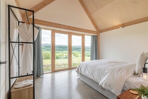 A bedroom with a large bed, a wooden ceiling, a window with a view of greenery, and a robe and slippers on a shelf.