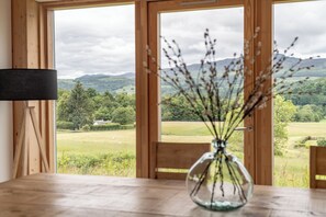 A wooden table with a glass vase containing dried plants, a wooden chair, and a view of a green landscape through large glass doors.
