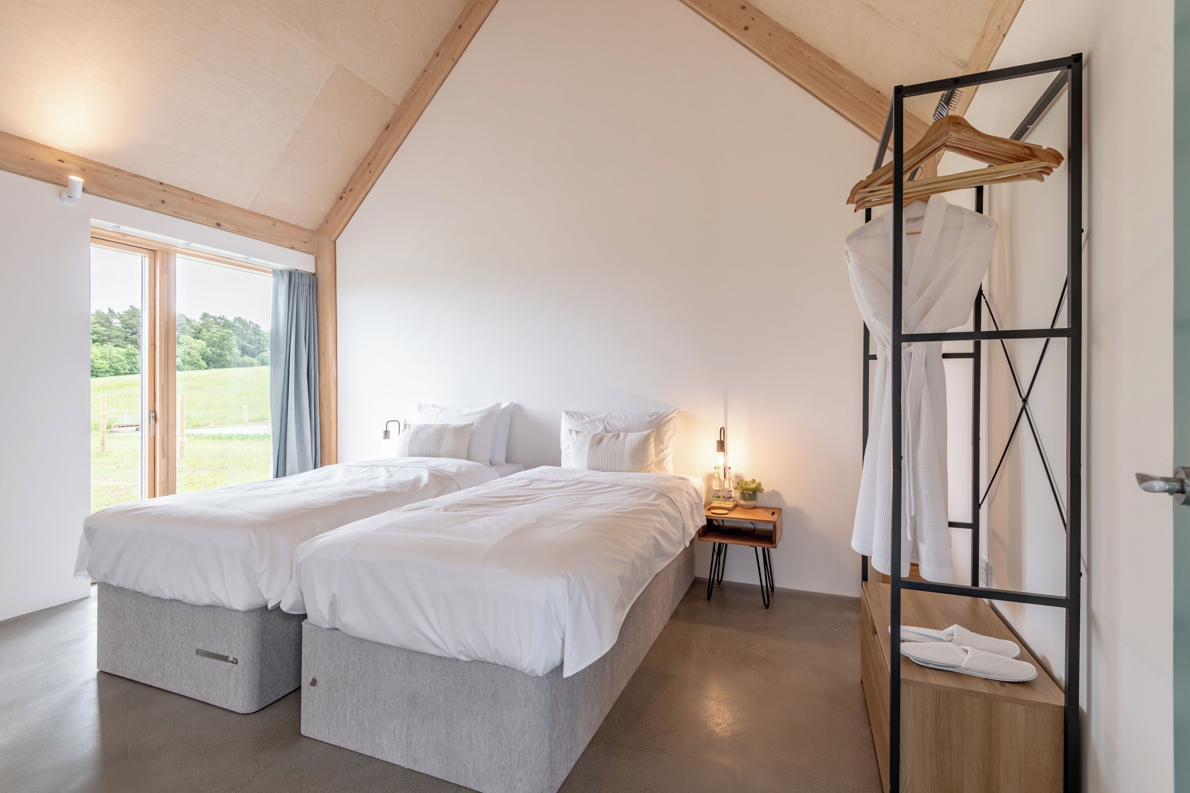 A bedroom with two beds, a wooden ceiling, a window with a view, and a hanging clothes rack.