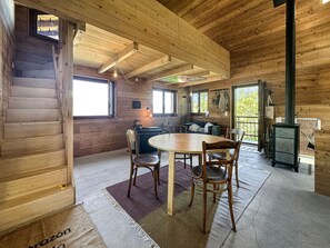 A wooden cabin interior with a dining area, a sofa, a fireplace, and a window overlooking greenery. - Alpine N'est Chalet - Stunning Mountain Views (Les Agettes)