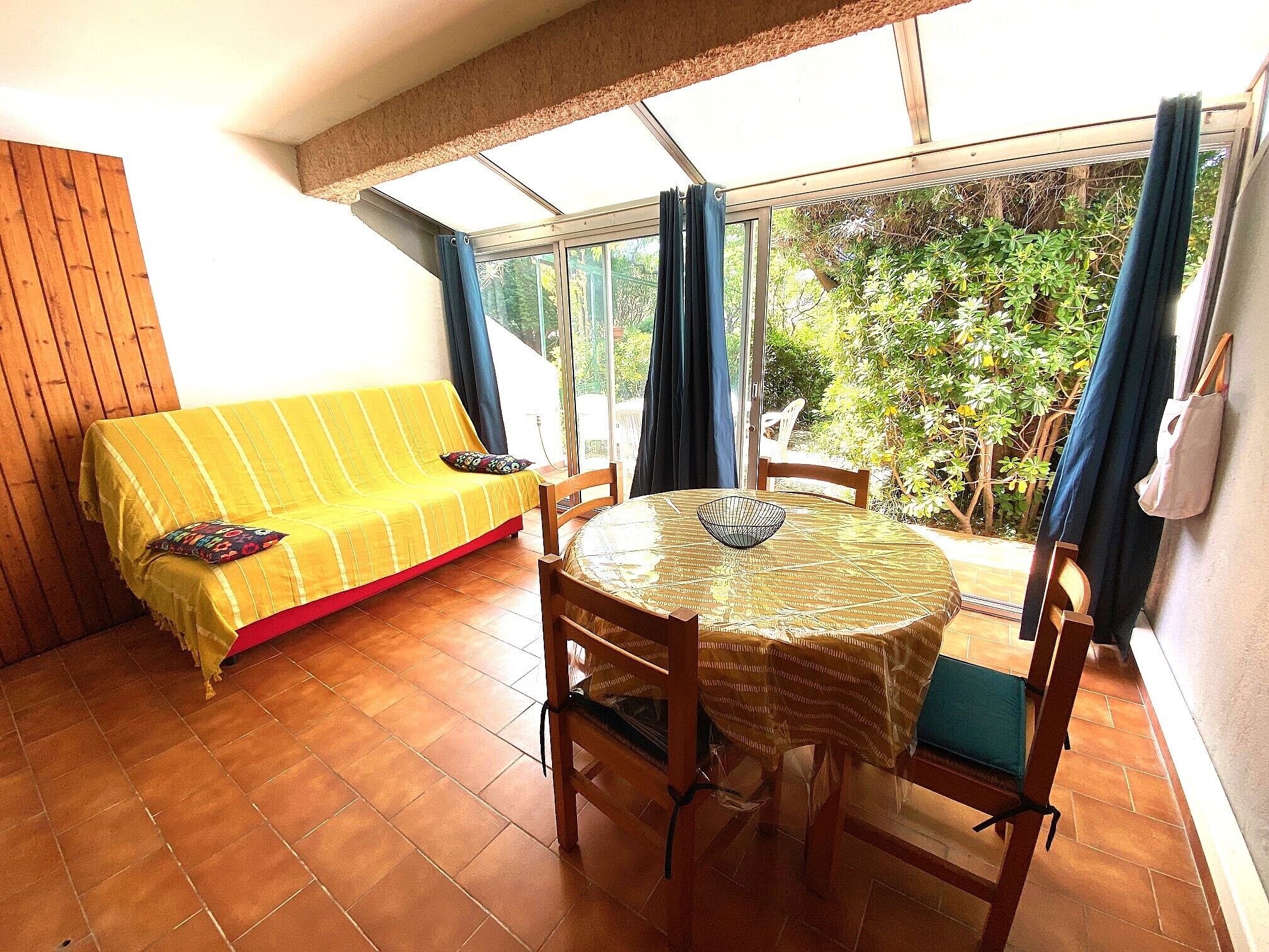 Ein Zimmer mit einem gelben Sofa, einem runden Tisch mit Stühlen und Blick auf Grün hinter großen Fenstern.