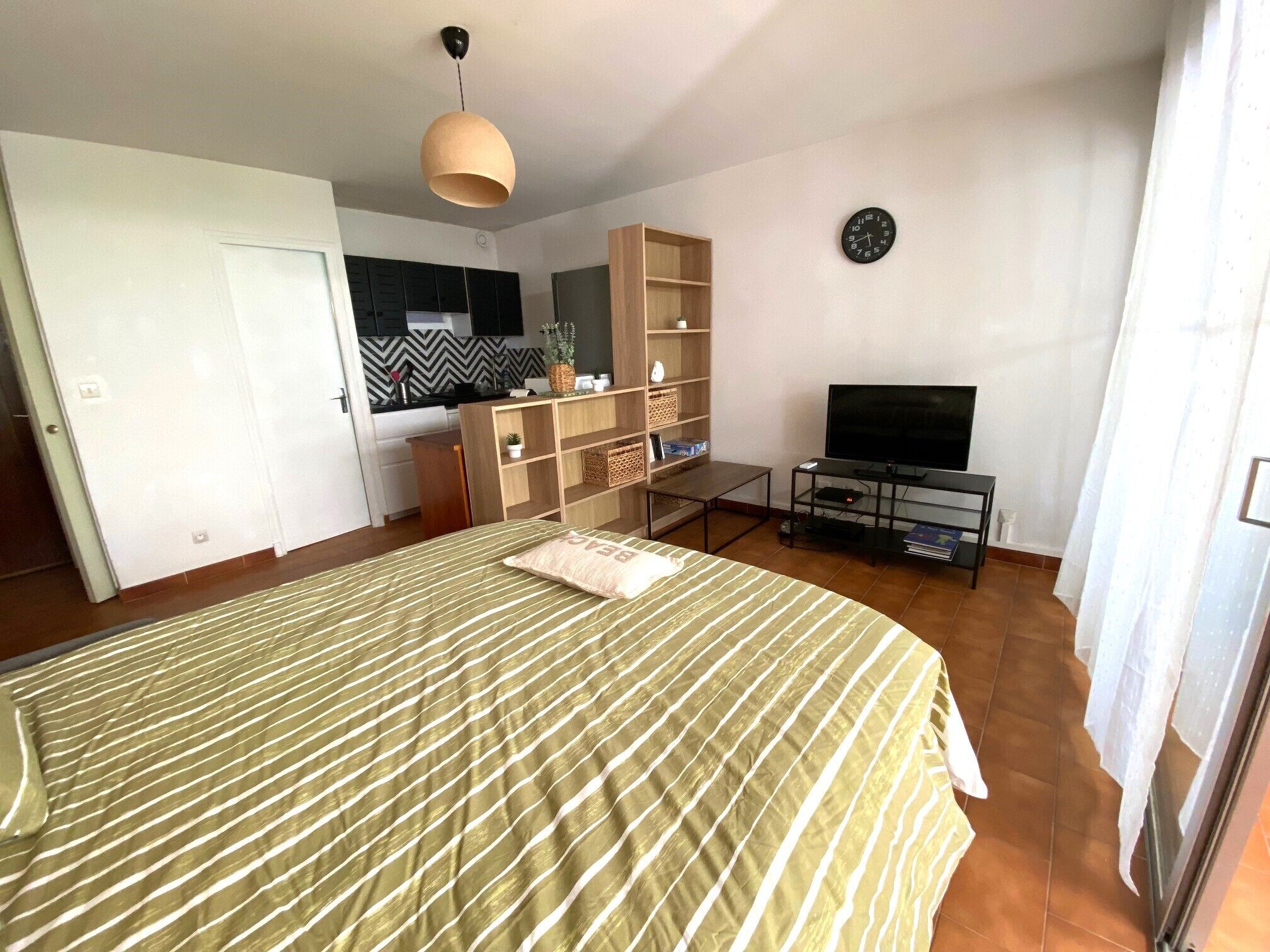 A bedroom with a bed, a TV, a small table, and a kitchenette in the background.