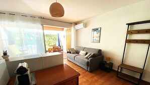 A living room with a sofa, a small table, a lamp, and a window with curtains. - "Le Maddy" studio garden-wifi-climate-parking-beach side (Saint-Cyr-sur-Mer)