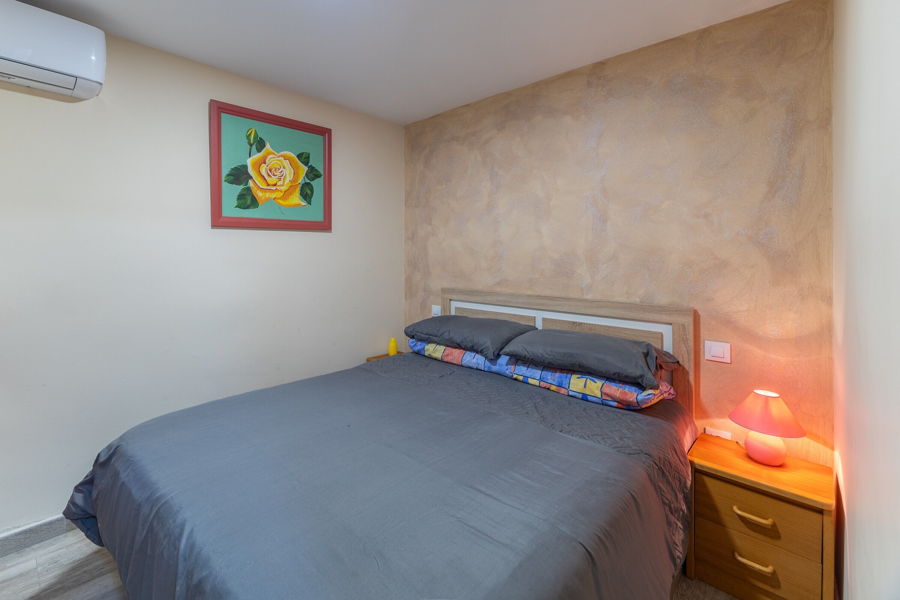Un dormitorio con cama, mesita de noche con lámpara, aire acondicionado de pared y un cuadro con una rosa.
