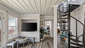A modern living room with a flat-screen TV, a small round table, a bench, a staircase, and a dining area in the background. - Beachfront | 3BR | Deck + Gulf Views (Galveston)