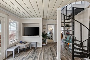 A modern living room with a flat-screen TV, a small round table, a bench, a staircase, and a dining area in the background. - Beachfront | 3BR | Deck + Gulf Views (Galveston)