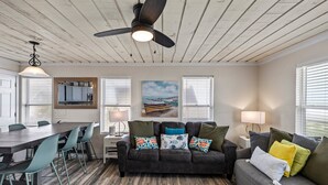A room with a wooden ceiling, a ceiling fan, a dining table with chairs, a sofa with cushions, and a painting on the wall. - Beachfront | 3BR | Deck + Gulf Views (Galveston)
