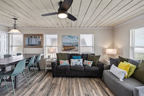 A room with a wooden ceiling, a ceiling fan, a dining table with chairs, a sofa with cushions, and a painting on the wall. - Beachfront | 3BR | Deck + Gulf Views (Galveston)