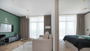 Una habitación de hotel moderna con sofá, una mesita redonda, una cama con colcha negra, un televisor montado en una pared verde y un ventanal con cortinas.